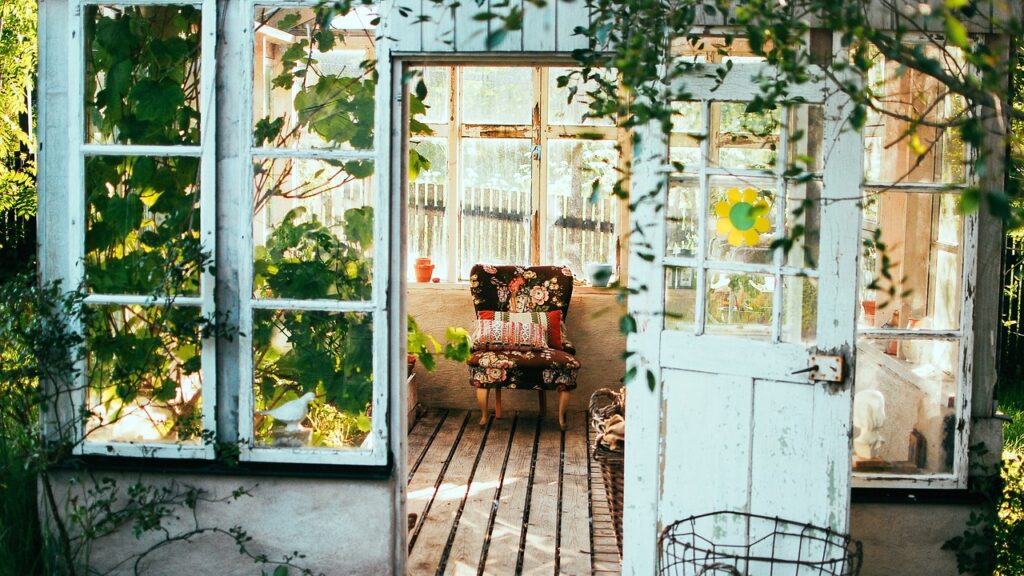 a shot from outsite a living room with windows and white paint as green plants hand over the door. inside we can see some furniture and vinyl flooring